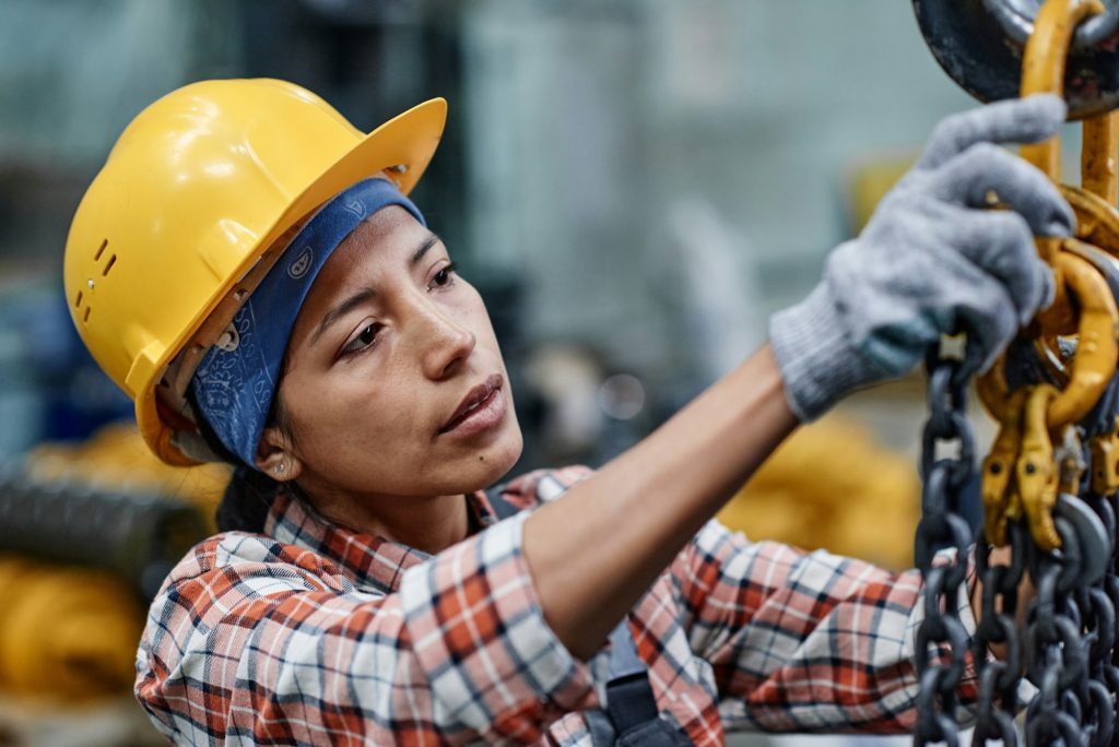 Young Hispanic female engineer in hardhat and gloves checking chains of machine